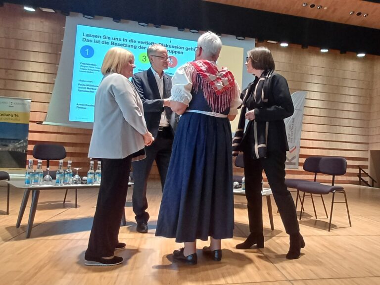 Vier Personen stehen im Kreis auf einer Bühne und besprechen sich. Im Hintergrund sieht man eine Leinwand mit der Projektion von Gruppeneinteilungen.