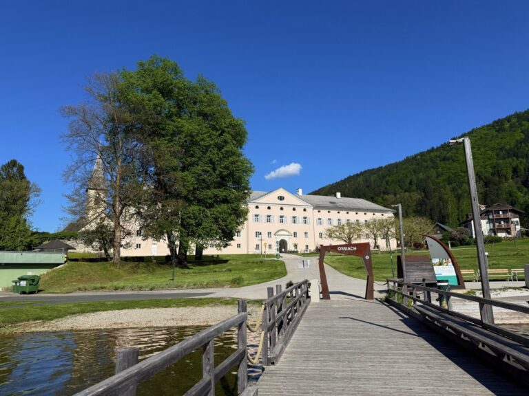 Außenansicht von CMA Ossiach, der Bick geht über eine Brücke auf die Zufahrt zu einem kleinen Eingangsbogen des leicht rosafarbenen Gebäudes.