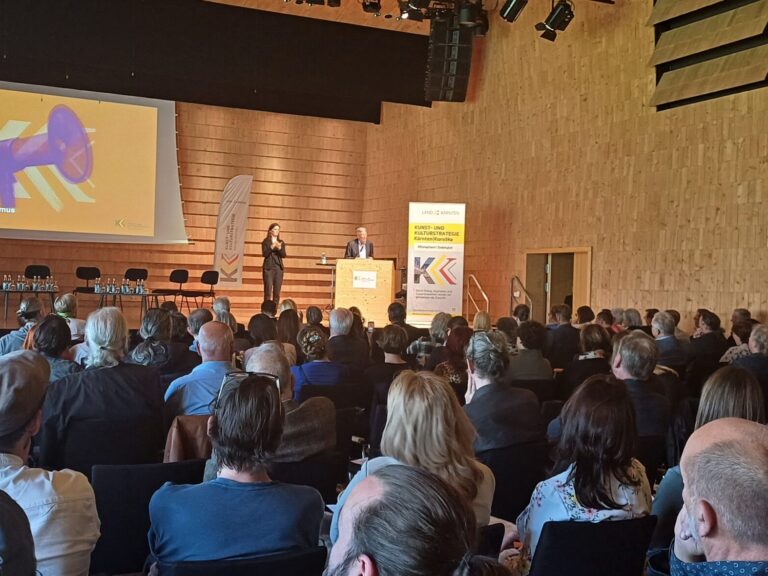 Im Vordergrund des Bildes sind die Köpfe des Publikums zu sehen, sie blicken auf die Bühne, dort steht HLH am Rednerpult, daneben die Gebärdendolmetscherin. Auf der Leinwand ist das Logo der Veranstaltung zu sehen, auf der Bühne sind außerdem Fahnen und Aufsteller der Kunst- und Kulturstrategie.
