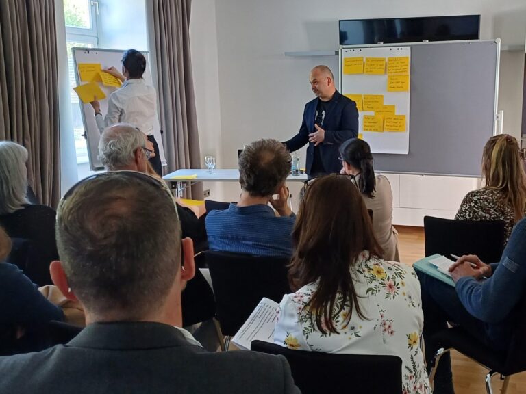 Eine Arbeitsgruppe ist von hinten zu sehen, der Blick geht auf den Moderator, der gestikuliert, eine weitere Person klebt Post-its auf eine Flip-Chart.