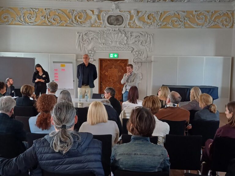 Eine Arbeitsgruppe ist von Hinten zu sehen, der Blick geht auf die Stirnseite des Raums, hier sind auf einer Flip-Chart Post-its zu erkennen, die Wand der Stirnseite ist mit Stuck verziert.