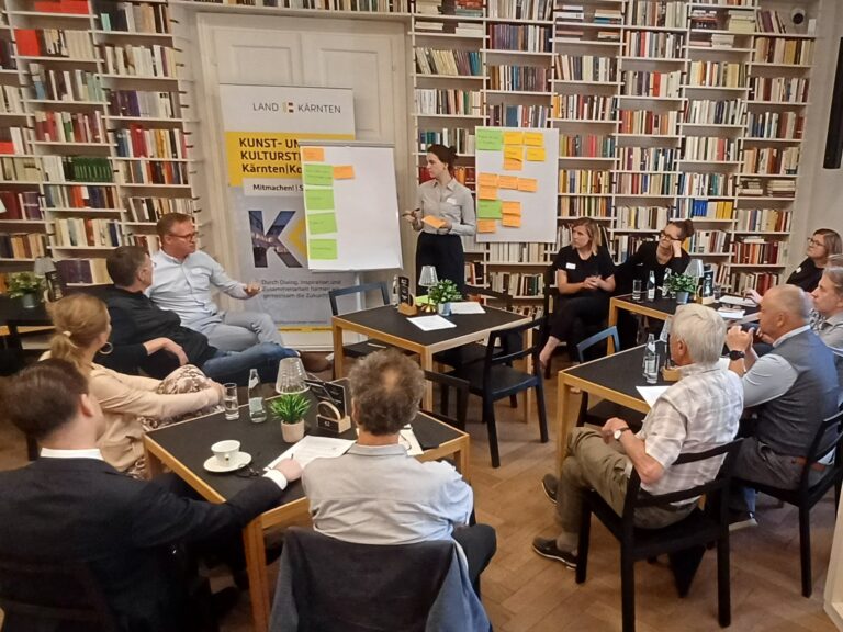Zwölf Personen sitzen in einem Halbkreis verteilt in einem Café Setting, die Blicke gehen auf zwei Flip-Chart Papiere, die mit Post-its beklebt sind, die hintere Wand besteht aus einem deckenhohen Bücherregal.