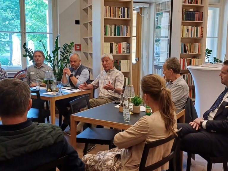 Eine Gruppe Workshop-Teilnehmende sitzen im Museumscafé, ein Teilnehmer spricht, im Hintergrund sind Bücher und ein Fenster.