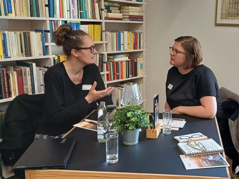 Zwei Frauen im Gespräch an einem Tisch, beide sind schwarz gekleidet und tragen eine dunkel umrahmte Brille, die linke Person trägt braune Haare im Dutt, die rechte Person hat kinnlanges hellbraunes Haar, im Hintergrund sind Bücher.