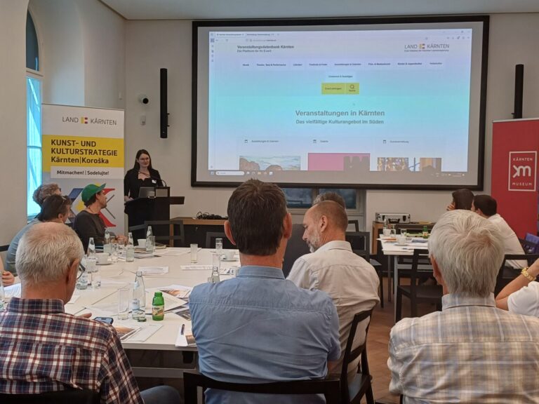 Workshop-Teilnehmende sind von Hinten sichtbar, der Blick geht auf eine Leinwand, vorne links spricht eine Referentin zum Veranstaltungsdatenraum Kärnten. Rechts im Bild sind Flip-Charts mit Post-its.