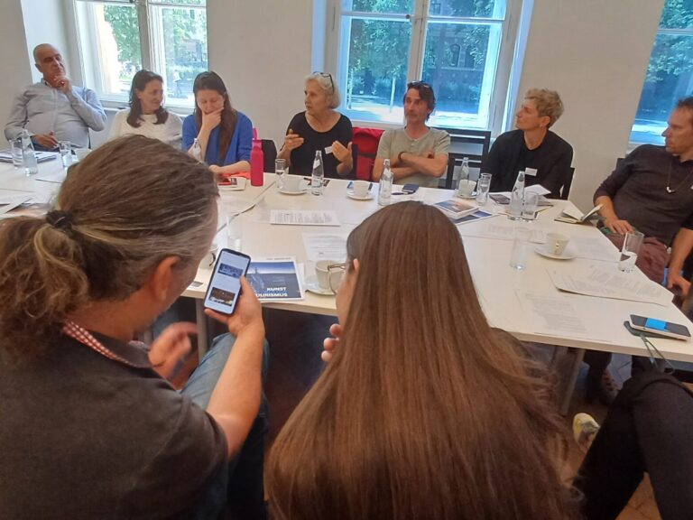 Workshop-Teilnehmende Sitzen um einen großen Tisch, im Vordergrund sind ein Mann mit langem Haar und eine Frau mit sehr langem Haar von hinten zu sehen, einander zugeneigt schauen sie beide auf dasselbe Handy.