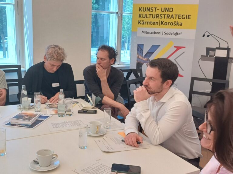Workshop-Teilnehmende sitzen am Tisch, rechts im Bild steht eine in schwarz gekleidete Frau mit Brille und gestikuliert.