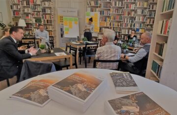 Im Vordergrund liegen Broschüren "A Cultural Journey" auf einem runden Stehtisch, im Hintergrund sind Workshop-Teilnehmende zu sehen, die rückseitige Wand besteht aus einem deckenhohen Bücherregal.