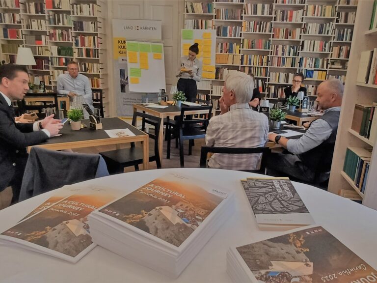 Im Vordergrund liegen Broschüren "A Cultural Journey" auf einem runden Stehtisch, im Hintergrund sind Workshop-Teilnehmende zu sehen, die rückseitige Wand besteht aus einem deckenhohen Bücherregal.