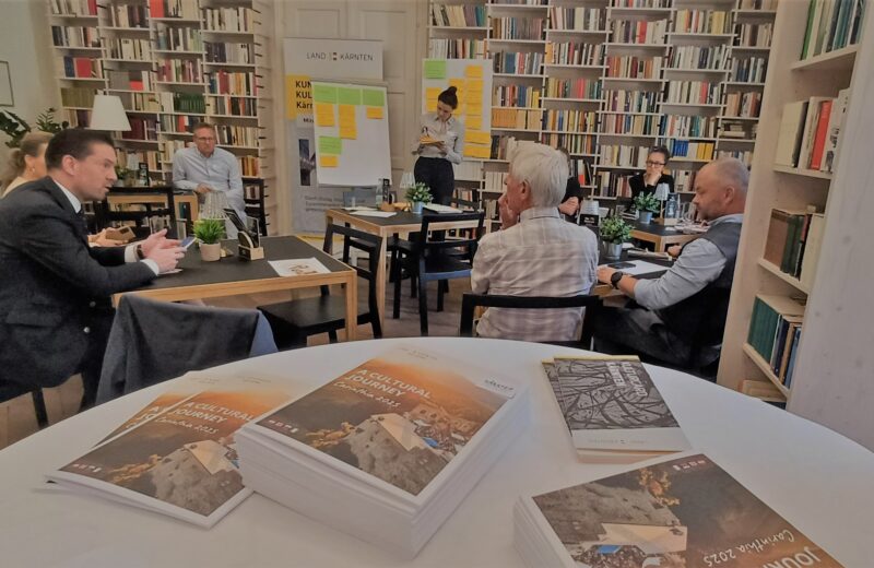 Im Vordergrund liegen Broschüren "A Cultural Journey" auf einem runden Stehtisch, im Hintergrund sind Workshop-Teilnehmende zu sehen, die rückseitige Wand besteht aus einem deckenhohen Bücherregal.