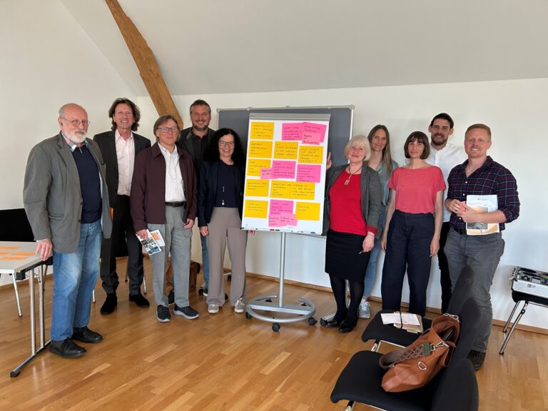Zehn Personen stehen rechts und Links von einer Flip-Chart, die mit gelben und pinken Post-its beklebt ist.