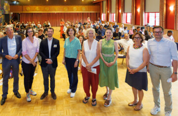 Im Vordergrund stehen acht Personen in einer gebogenen Linie, es handelt sich um die Impulsgeberinnen, Panel-Diskutant:innen, den Herrn Landeshauptmann Dr. Peter Kaiser und Frau Brigitte Winkler-Komar. Im Hintergrund ist ein Veranstaltungssaal zu sehen, das Ambiente ist von honigfarbenem Holz und roten Wänden und Vorhängen geprägt. Auf den Stühlen, mit Blick richtig Kamera sitzen die Teilnehmenden des Zukunftsforums.