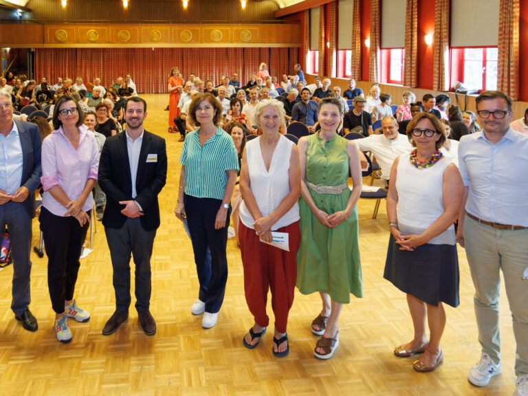 Im Vordergrund stehen acht Personen in einer gebogenen Linie, es handelt sich um die Impulsgeberinnen, Panel-Diskutant:innen, den Herrn Landeshauptmann Dr. Peter Kaiser und Frau Brigitte Winkler-Komar. Im Hintergrund ist ein Veranstaltungssaal zu sehen, das Ambiente ist von honigfarbenem Holz und roten Wänden und Vorhängen geprägt. Auf den Stühlen, mit Blick richtig Kamera sitzen die Teilnehmenden des Zukunftsforums.