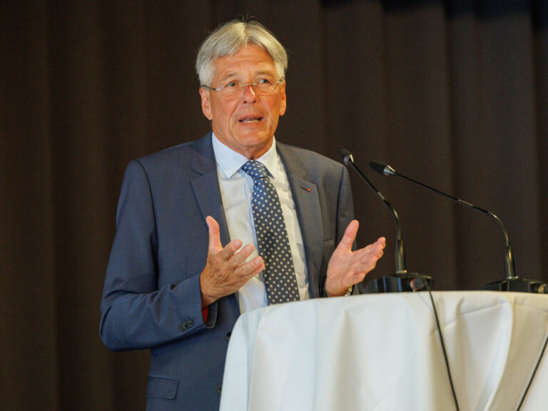 Vor einem schwarzen Vorhang steht Dr. Peter Kaiser hinter einem runden Tisch mit Tischdecke, darauf zwei Tischmikrofone. Er trägt einen blaugrauen Anzug, eine Krawatte in kleinem Schachbrettmuster und gestikuliert mit beiden Händen.