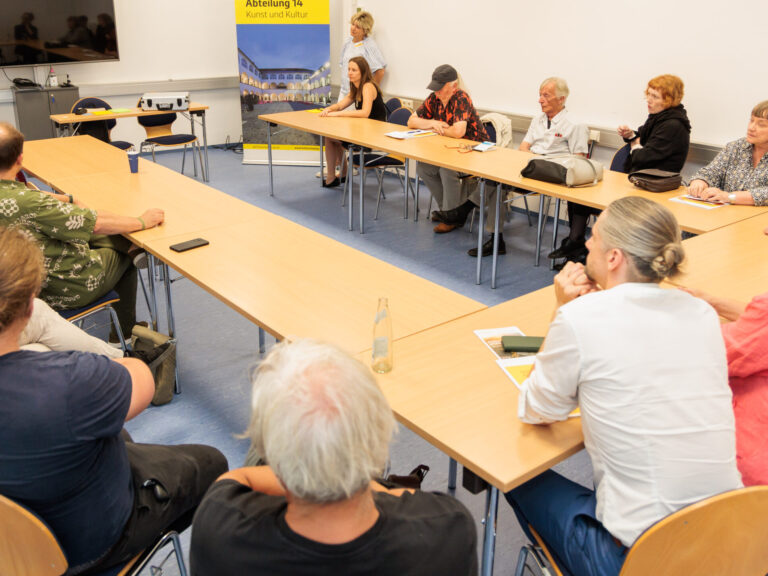 13 Teilnehmende einer Arbeitsgruppe sitzen an Tischen in U-Formation und Blicken auf Angelika Hödl, oben links im Bild.
