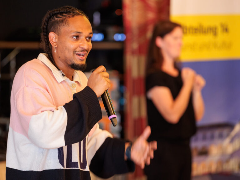 Der Rapper Evan Parks ist im Vordergrund zu sehen, sein Blick geht nach rechts, in einer Hand hält er ein Mikrofon. Er trägt eine Flechtfrisur, einen silbernen Ohrring und einen Pulli mit sehr breiten Streifen: Oben Rosa, Mitte Weiß, unten Dunkelblau/schwarz. Im Hintergrund ist verschwommen eine Gebärdendolmetscherin zu sehen.