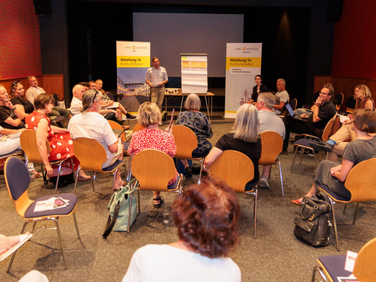 Teilnehmende einer Arbeitsgruppe sitzen in zwei Reihen im Halbkreis, die Blicke auf den Moderator, eine Flip-Chart und zwei Aufsteller ausgerichtet.