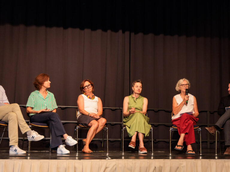 Vor einem schwarzen Vorhand sitzen die Diskutant:innen der Panel-Diskussion im Halbkreis. Von links nach Rechts: Michael Aichholzer, Claudia Ströbitzer, Gabriele Semmelrock-Werzer, Ulrike Kuner, Angelika Hödl (sie hält ein Mikrofon) und der Moderator Sebastian Lücke.