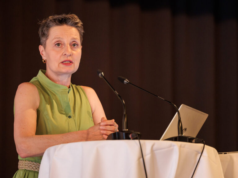Ulrike Kuner steht am Rednerpult und sprich in ein Tisch-Mikrofon. Ihr Blick geht knapp am Betrachtenden vorbei. Sie trägt einen Kurzhaarschnitt, kleine goldene Kreolenohrringe und ein grünes Kleid.