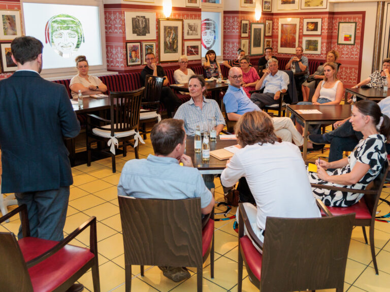 Teilnehmende einer Arbeitsgruppe sitzen im Café des Fuchspalasts an kleinen Tischen verteilt, die Blicke gehen überwiegend nach links zu den zwei Moderator:innen. Das Café hat einen gelben Fliesenboden, dunkles Holzmobiliar und rot-weiß gemusterte Wände mit gerahmten Bildern.