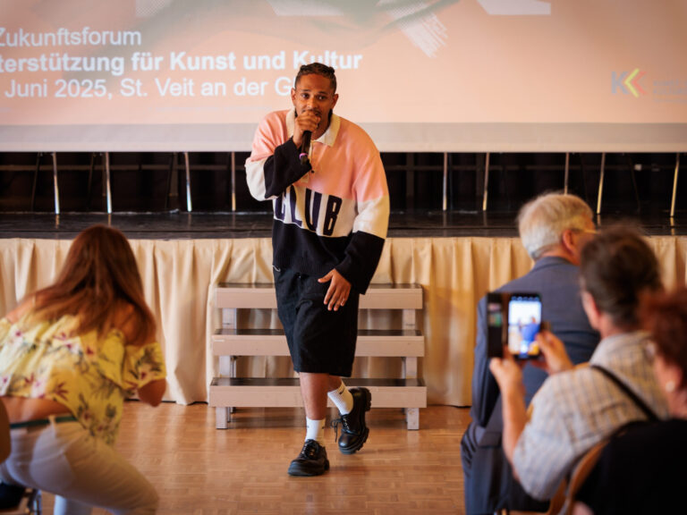 Der Rapper Evan Parks ist im Zentrum des Bildes, sein Blick geht nknapp am Betrachter vorbei, in einer Hand hält er ein Mikrofon. Er trägt eine Flechtfrisur, einen Pulli mit sehr breiten Streifen: Oben Rosa, Mitte Weiß, unten Dunkelblau/schwarz; eine schwarze Hose bis über die Knie, weiße Socken und klobige schwarze Schuhe. Rechts und links von ihm sind durch den Mittelgang Teilnehmende des Zukunftsforums von hinten und verschwommen sichtbar.
