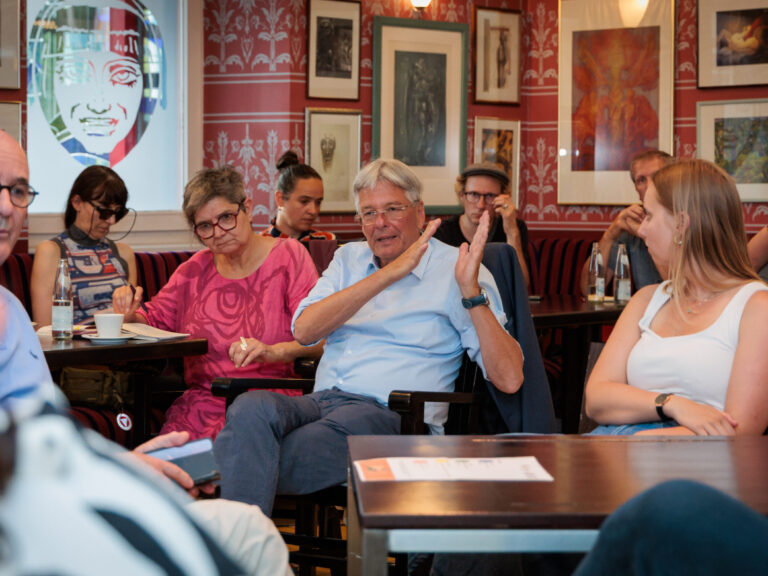In der Bildmitte sitzt Dr. Peter Kaiser im Café des Fuchspalasts, er spricht und gestikuliert. Um ihn herum sitzen weitere Teilnehmende der Arbeitsgruppe. Das Raumambiente ist von dunklen Holzmöbeln und rot-weiß gemusterten Wänden mit gerahmten Bildern geprägt.