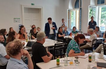 Der Blick geht in den Raum, die Workshopteilnehmenden sitzen an Tischgruppen verteilt, drei Personen stehen. Eine stehende Person in der Mitte trägt einen dunklen Sakko mit einem Namensschild, darunter ein weißes Hemd. Er blickt auf seine Notizen auf dem Tisch und spricht zu den Teilnehmenden.