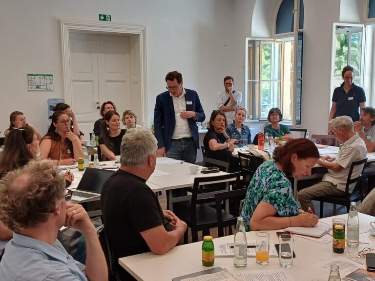 Der Blick geht in den Raum, die Workshopteilnehmenden sitzen an Tischgruppen verteilt, drei Personen stehen. Eine stehende Person in der Mitte trägt einen dunklen Sakko mit einem Namensschild, darunter ein weißes Hemd. Er blickt auf seine Notizen auf dem Tisch und spricht zu den Teilnehmenden.