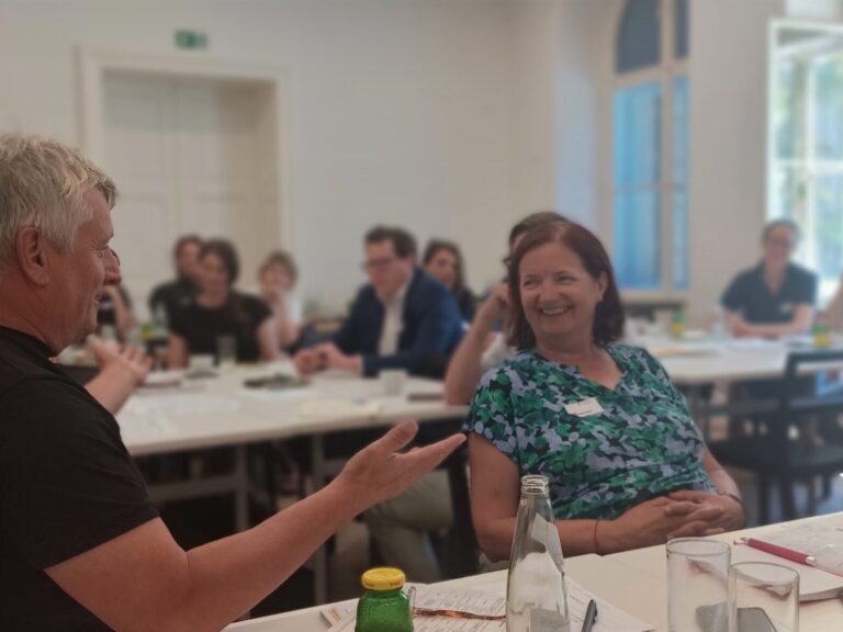 Zwei Teilnehmende der Werkstatt sitzen einander zugewandt, rechts ist das lachende Gesicht einer Frau mit blau gemustertem T-Shirt nach links gedreht, sie schaut einen Mann mit kurzem grauen Haar an, er ist schräg von hinten zu sehen macht mit beiden Armen eine einladende Geste.