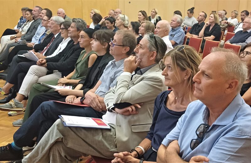 Ein Konferenzraum mit mehreren Reihen von sitzenden Personen, die vermutlich aufmerksam einer Präsentation folgen; einige halten Unterlagen in den Händen.