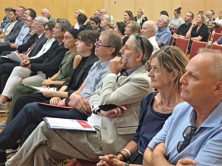 Ein Konferenzraum mit mehreren Reihen von sitzenden Personen, die vermutlich aufmerksam einer Präsentation folgen; einige halten Unterlagen in den Händen.