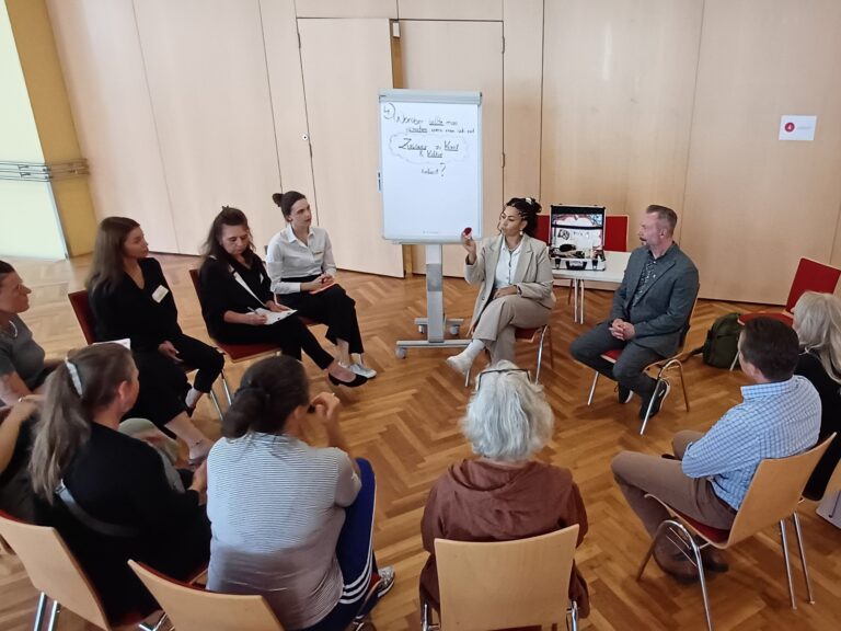 Zwölf Personen sitzen in einem Stuhlkreis in einem hölzern verkleideten Raum. Die Frau in der Bildmitte ist am Sprechen und scheint etwas in der Hand zu halten, links neben ihr ist ein Flipchart, auf dem schon etwas steht.