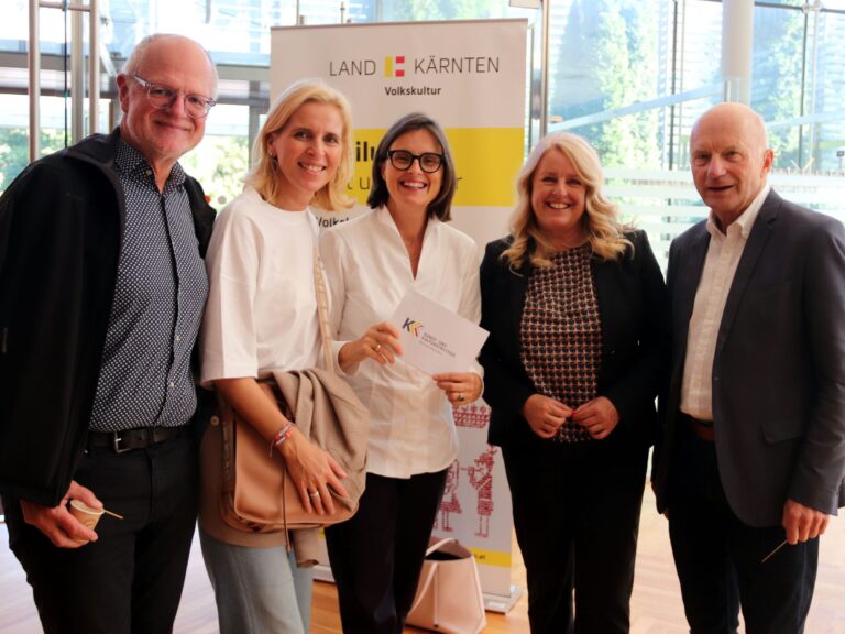 Fünf formell gekleidete Personen posieren lächelnd für ein Foto, die Frau in der Mitte hält Moderationskarten in den Händen; im Hintergrund ist ein Banner mit dem Schriftzug "Land Kärnten" zu sehen.