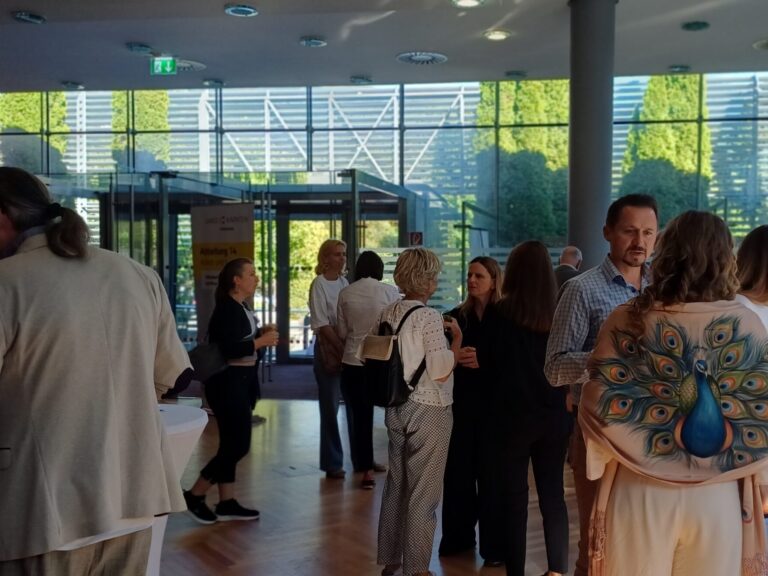 Personen stehen in einer modernen, lichtdurchfluteten Lobby, unterhalten sich und genießen Getränke an einem Buffet-Tisch mit Wasserflaschen und Snacks.