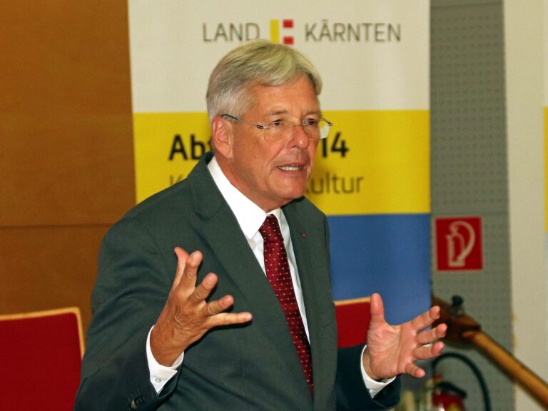 Kulturreferent und Landeshauptmann im Anzug, der anscheinend am Reden ist und mit den Händen gestikuliert, im Hintergrund ein Banner mit dem Schriftzug "Land Kärnten".