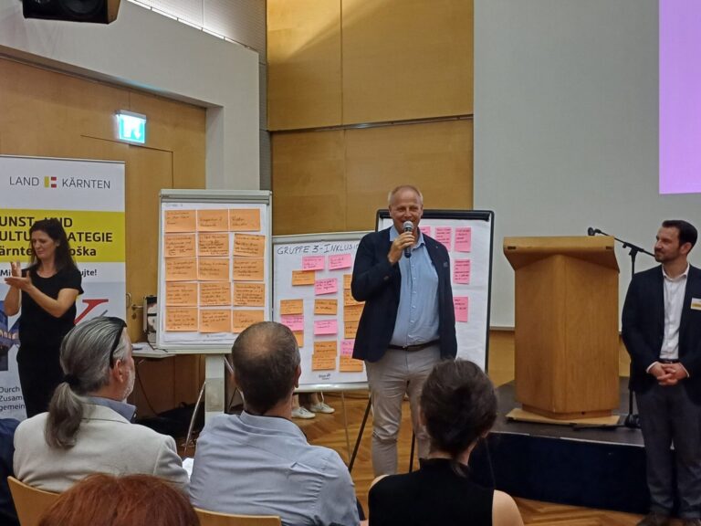Im Vordergrund Rücken von Menschen im Publikum, in der Mitte des Bildes eine Frau die mit ihren Händen gestikuliert und zwei Männer, einer mit einem Mikrofon in der Hand, der einen Vortrag hält; im Hintergrund befüllte Flipcharts; rechts ist auch ein Teil der Bühne zu sehen.