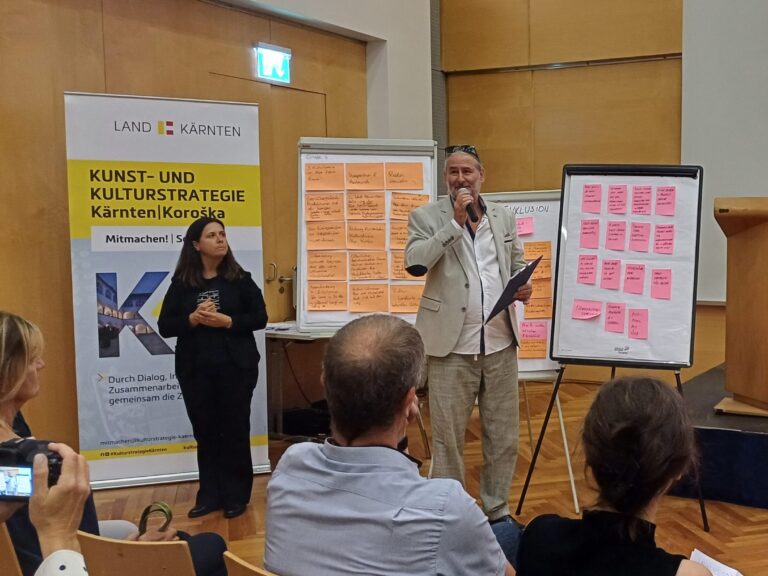 Im Vordergrund Rücken von Menschen im Publikum, in der Mitte des Bildes eine Frau und ein Mann mit einem Mikrofon in der Hand, der einen Vortrag hält; im Hintergrund befüllte Flipcharts.
