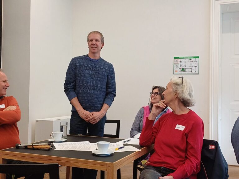 Ein Mann steht in einem hellen Raum neben einem Holztisch, umgeben von vier sitzenden Personen, die ihm zuhören. Auf dem Tisch liegen Dokumente, Stifte und Tassen. Die Personen schauen den Redner interessiert an.