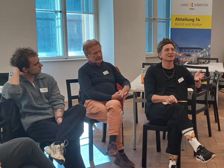 Drei Personen sitzen in einem hellen Raum auf einer Stuhlreihe und sprechen über ein Thema. Im Hintergrund sind Fenster zu sehen, die Tageslicht in den Raum lassen. Rechts im Hintergrund ein Banner der Abteilung 14 Kunst und Kultur.