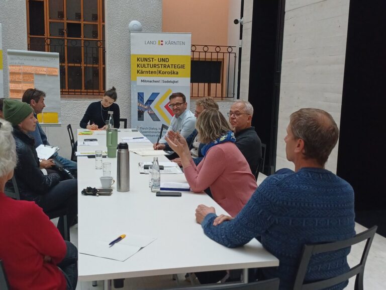 Seitliche Ansicht auf einen rechteckigen Tisch, an dem mehrere Personen sitzen. Eine Frau am Kopfende schreibt Notizen, während die anderen aktiv diskutieren. Im Hintergrund befinden sich zwei Banner der Abteilung 14 und ein beschriftetes Flipchart