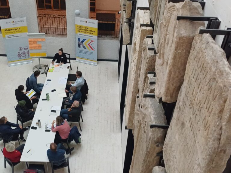 Vogelperspektive auf eine kleine Workshop-Gruppe, die an einem weißen Konferenztisch sitzt. Der rechte Bildrand zeigt mehrere vertikal aufgehängte Steinreliefs in einem modernen Ausstellungsraum. Im Hintergrund stehen Banner, auf einer Staffelei ist ein beschriftetes Flipchart zu sehen.