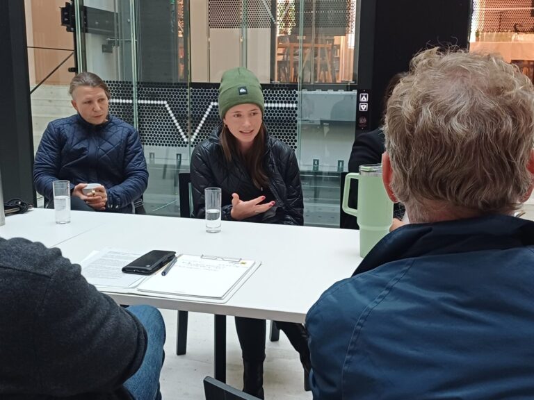 Mehrere Personen sitzen um einen großen rechteckigen Tisch in einem modernen und hellen Raum - dem Foyerbereich vom kärnten.museum. Eine Frau in einer dunklen Jacke spricht gestikulierend, während die anderen Personen interessiert zuhören. Auf dem Tisch liegen Notizzettel, Stifte und Wasserflaschen.