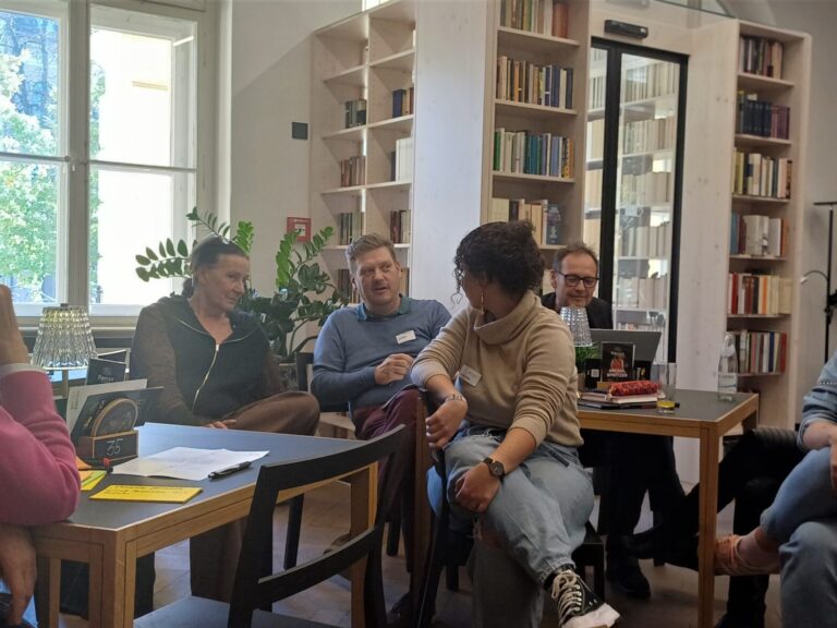 Eine Gruppe von sechs Personen sitzt in im Museums-Café um zwei Tische herum. Im Hintergrund sind mehrere Bücherregale und große Fenster zu sehen. Die Personen unterhalten sich und wirken konzentriert.