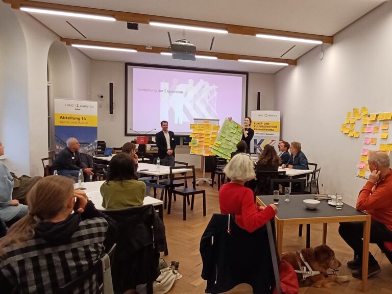 Ein mittelgroßer, heller Konferenzraum mit Bestuhlung in kleinen Gruppen. Im Bild sitzen mehrere Personen um Tische, die in Gruppen angeordnet sind. Eine Person steht vorne und präsentiert die Ergebnisse eines Workshops. Neben dieser Person sind bunte Moderationskarten auf großen Pinnwänden sichtbar. Im Hintergrund eine Leinwand mit einer Folie und Bannern.