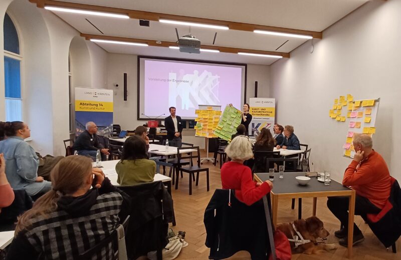 Ein mittelgroßer, heller Konferenzraum mit Bestuhlung in kleinen Gruppen. Im Bild sitzen mehrere Personen um Tische, die in Gruppen angeordnet sind. Eine Person steht vorne und präsentiert die Ergebnisse eines Workshops. Neben dieser Person sind bunte Moderationskarten auf großen Pinnwänden sichtbar. Im Hintergrund eine Leinwand mit einer Folie und Bannern.